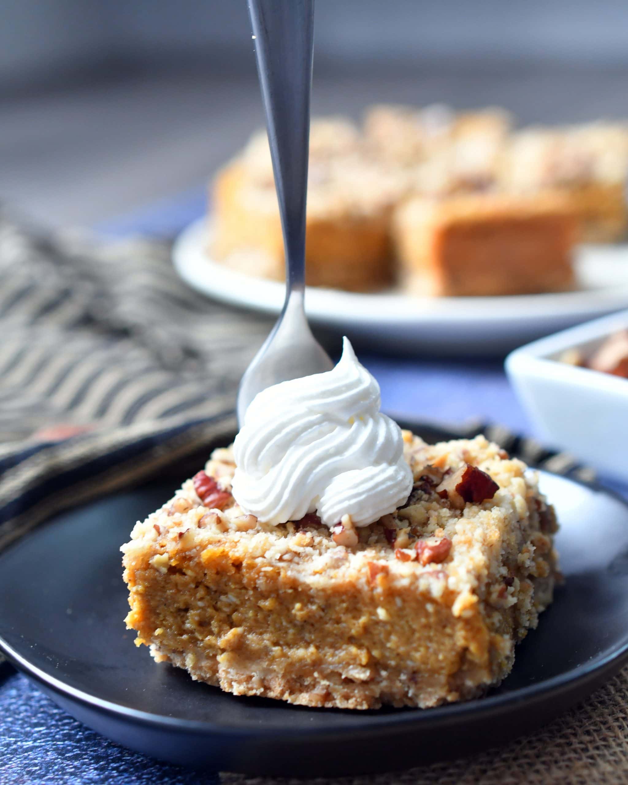 pecan pumpkin cheesecake bar topped with whipped cream, fork inserted in bar, on a brown plate with additional bars in the background