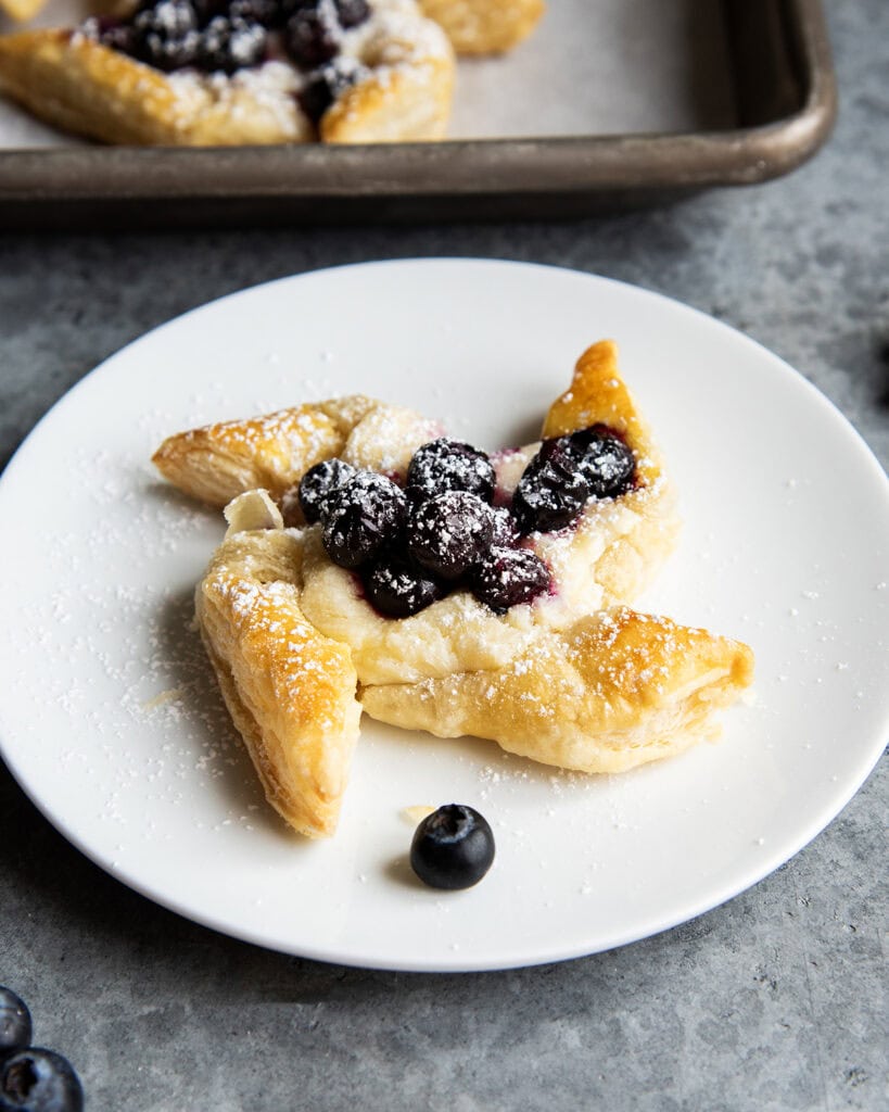 A pinwheel shaped danish topped with cream cheese, blueberries, and powdered sugar on a plate.