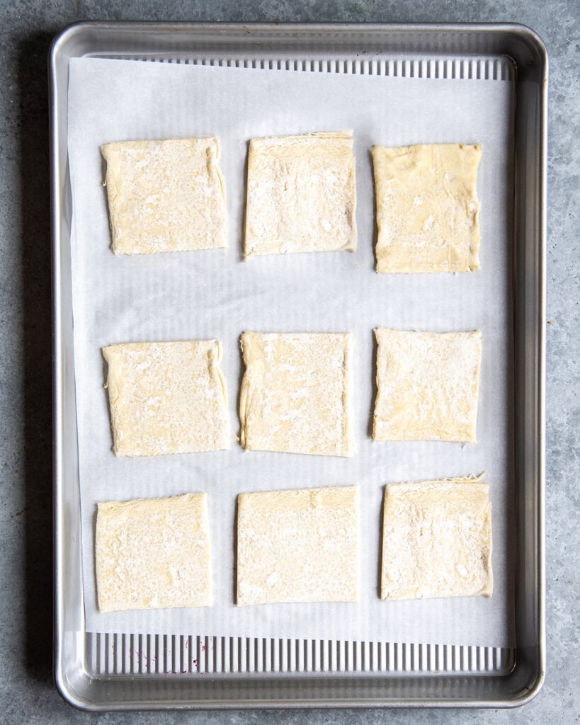 9 squares of raw puff pastry dough on a cookie sheet.