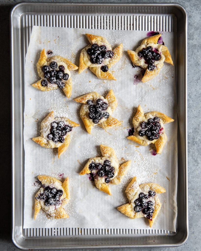An above photo of Blueberry Danishes in the shape of pinwheels on a baking sheet.