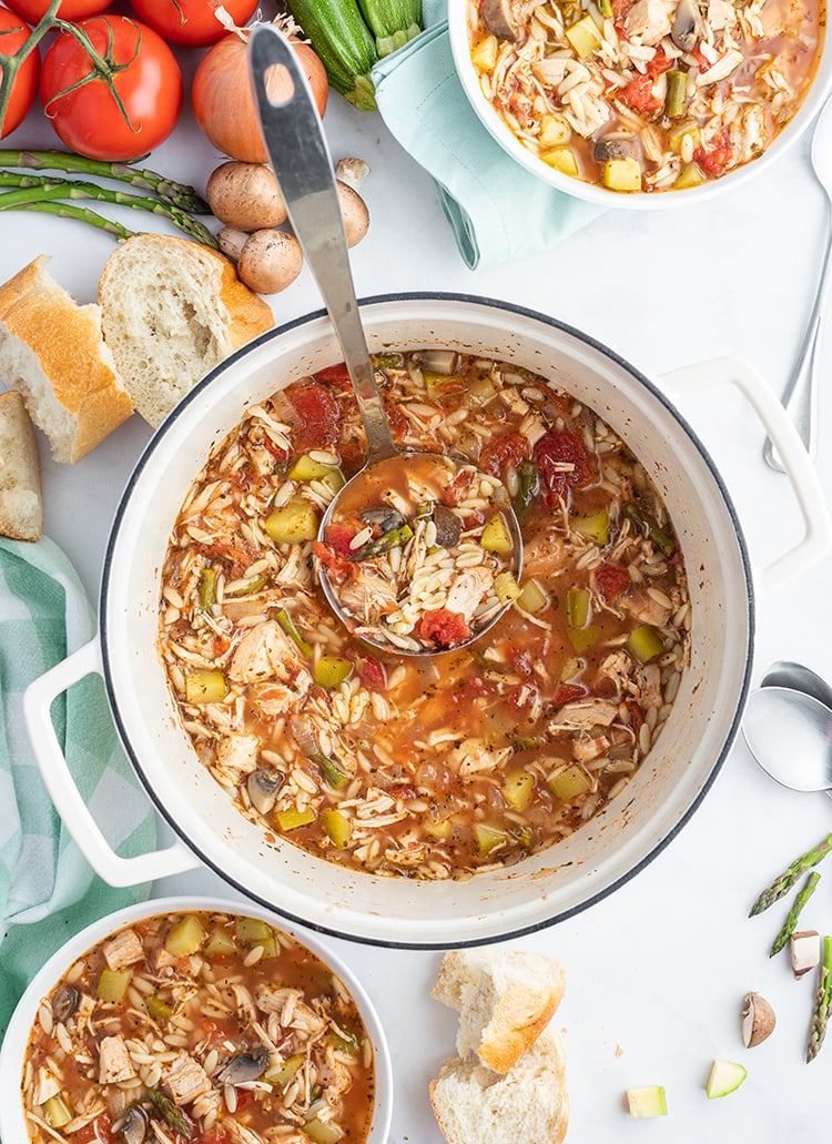 A pot of Italian Turkey Orzo Soup with two bowls of soup on the side and ingredients of the soup next to it.