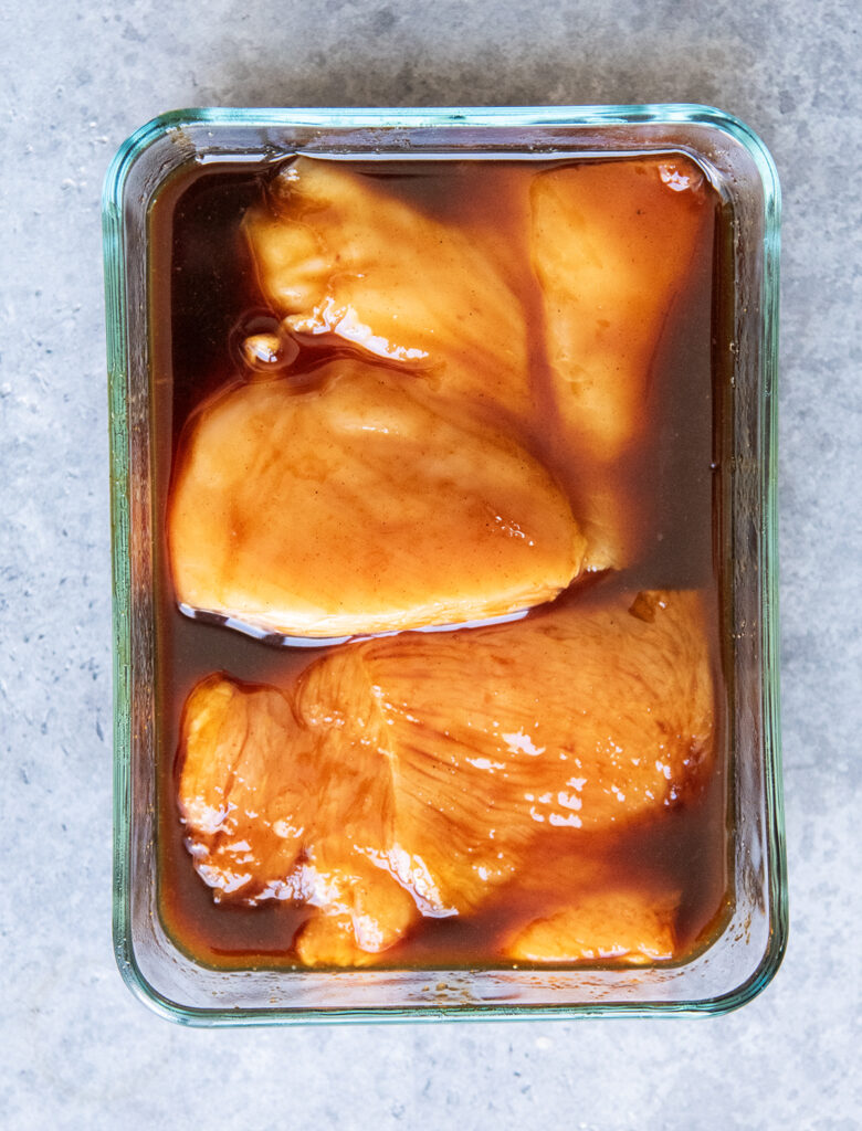 A glass container with chicken breasts marinating in teriyaki sauce. 