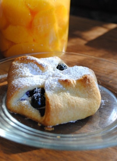 Angled view of blueberry cream cheese danish on a glass plate with peaches in background.