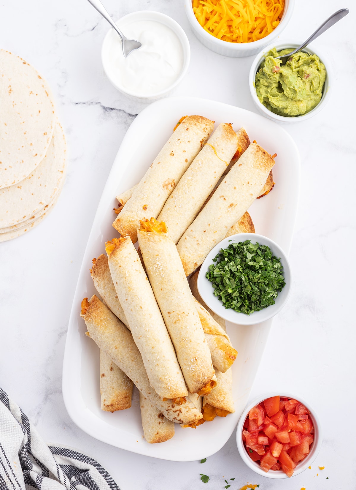 An overhead photo of taquitos on a serving plate surrounded by cilantro, diced tomatoes, and guacamole.