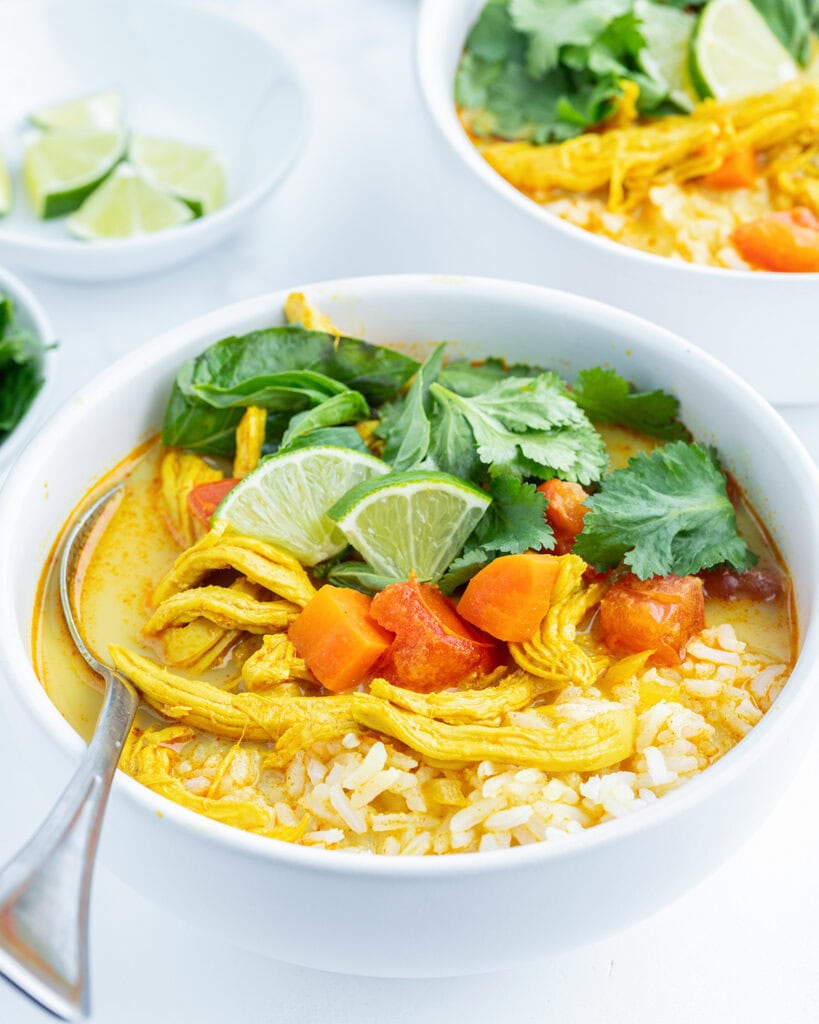 A close up of a bowl of chicken curry soup with rice, carrots, and topped with lime wedges, and fresh herbs.