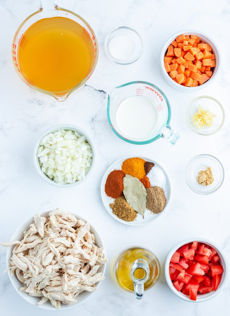 The ingredients needed to make chicken curry soup, like chicken broth, carrots, chicken, tomato, and a plate of spices. 
