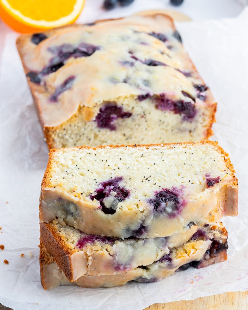 A loaf of blueberry poppy seed bread with some sliced in front and laying on a piece of parchment paper.