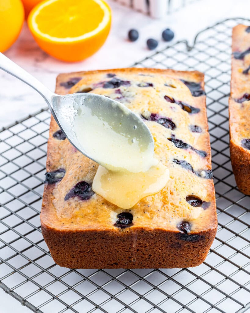 A spoon topping a loaf of blueberry poppyseed bread with orange glaze.
