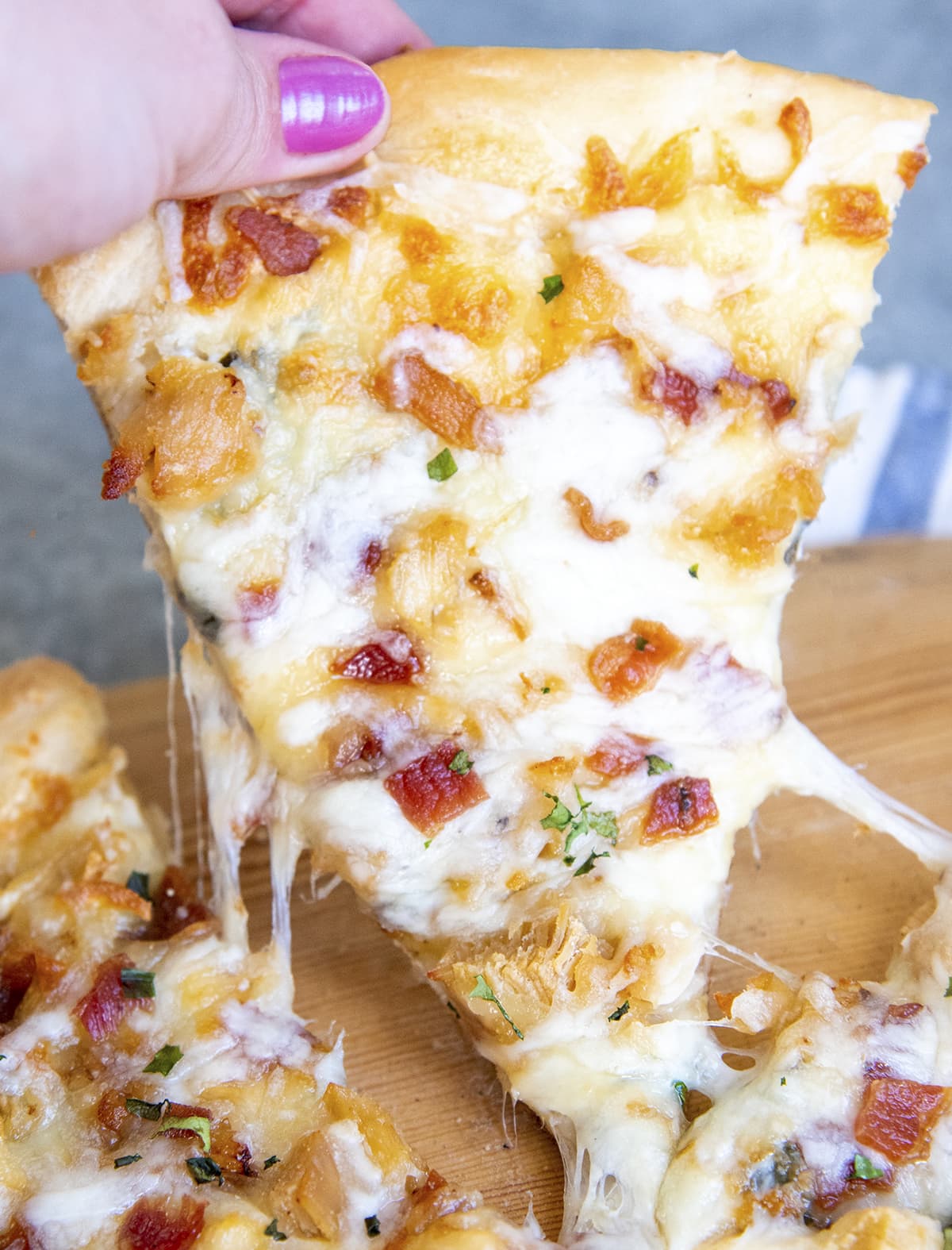 A hand holding a piece of chicken, bacon, alfredo pizza.