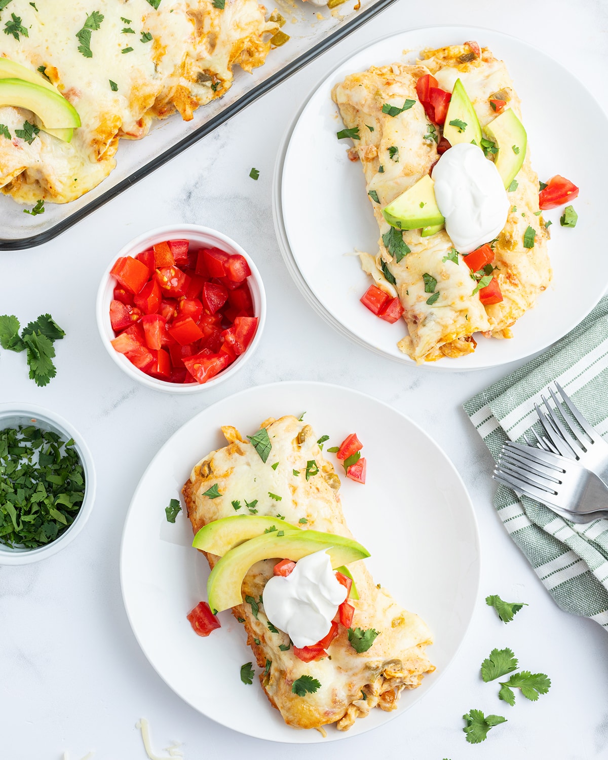 An overhead photo of two plates of creamy chicken enchiladas topped with avocado slices, and a dollop of sour cream.