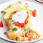 A close up of a plate of two chicken enchiladas topped with sour cream, tomatoes, and slices of avocado.