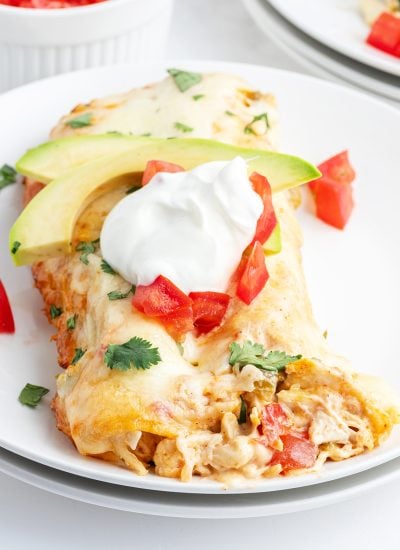 A close up of a plate of two chicken enchiladas topped with sour cream, tomatoes, and slices of avocado.