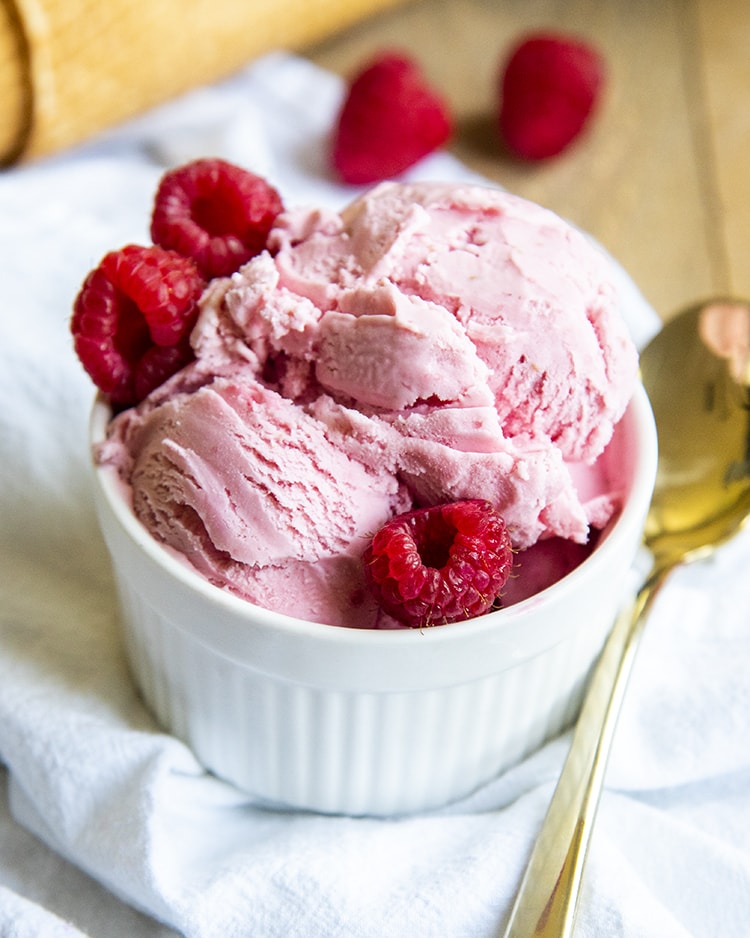 A small ramekin bowl filled with pink raspberry ice cream and surrounded by three raspberries.