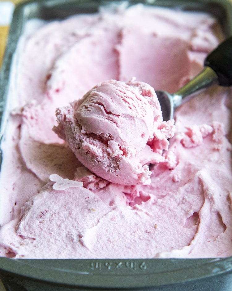 A scoop of pink ice cream in an ice cream scoop in a metal pan of pink ice cream.