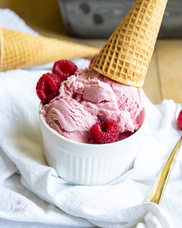 A small ramekin bowl filled with pink raspberry ice cream and surrounded by three raspberries, and topped with an upside down ice cream cone.