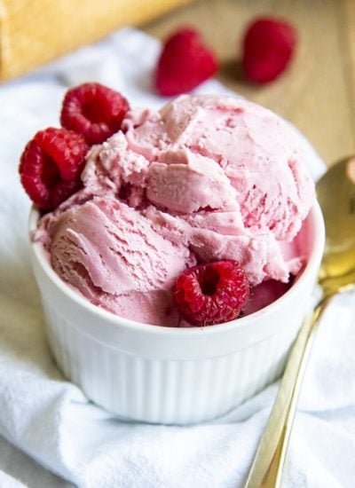 A small ramekin bowl filled with pink raspberry ice cream and surrounded by three raspberries.