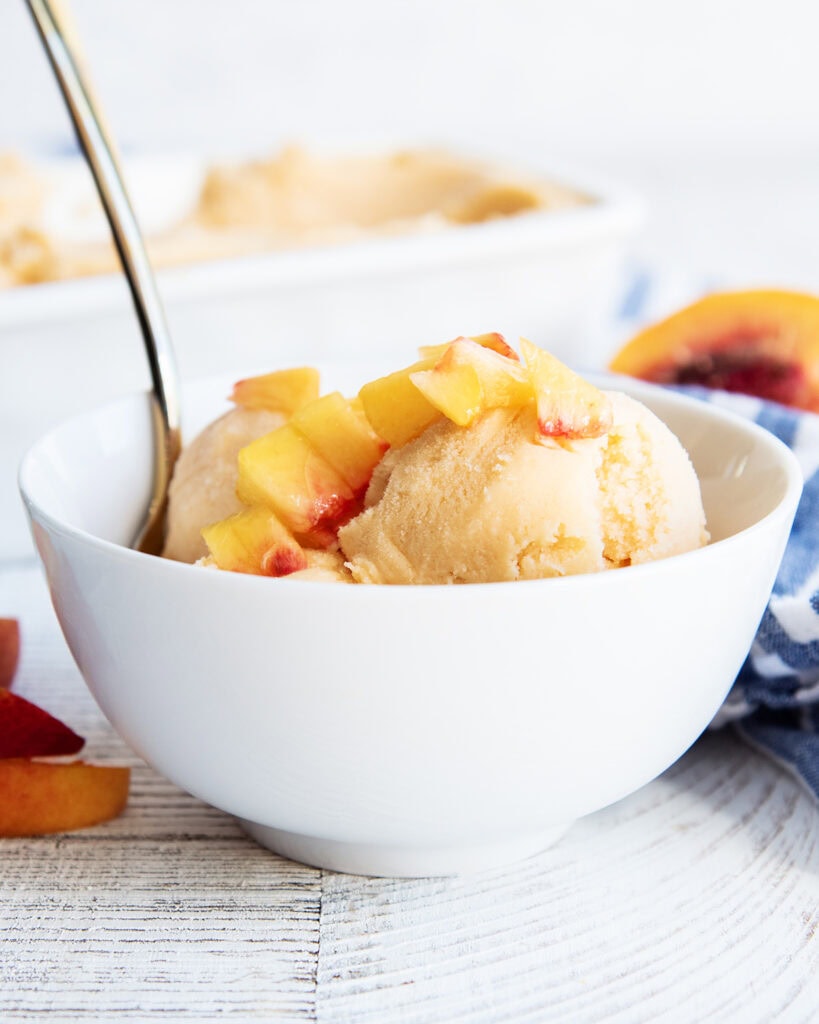 A bowl of peach sherbet topped with fresh diced peaches, with a spoon in it.