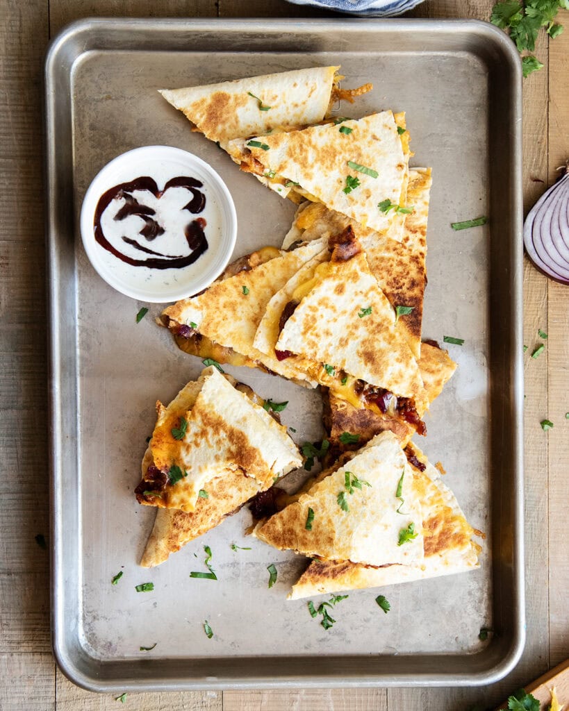 Slices of bbq chicken quesadilla on a metal pan next to a bowl of ranch dip.