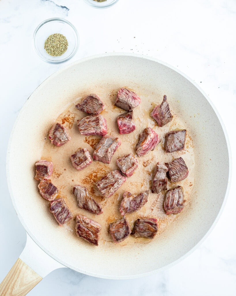 Beef stew meat pieces being browned in a white saute pan.