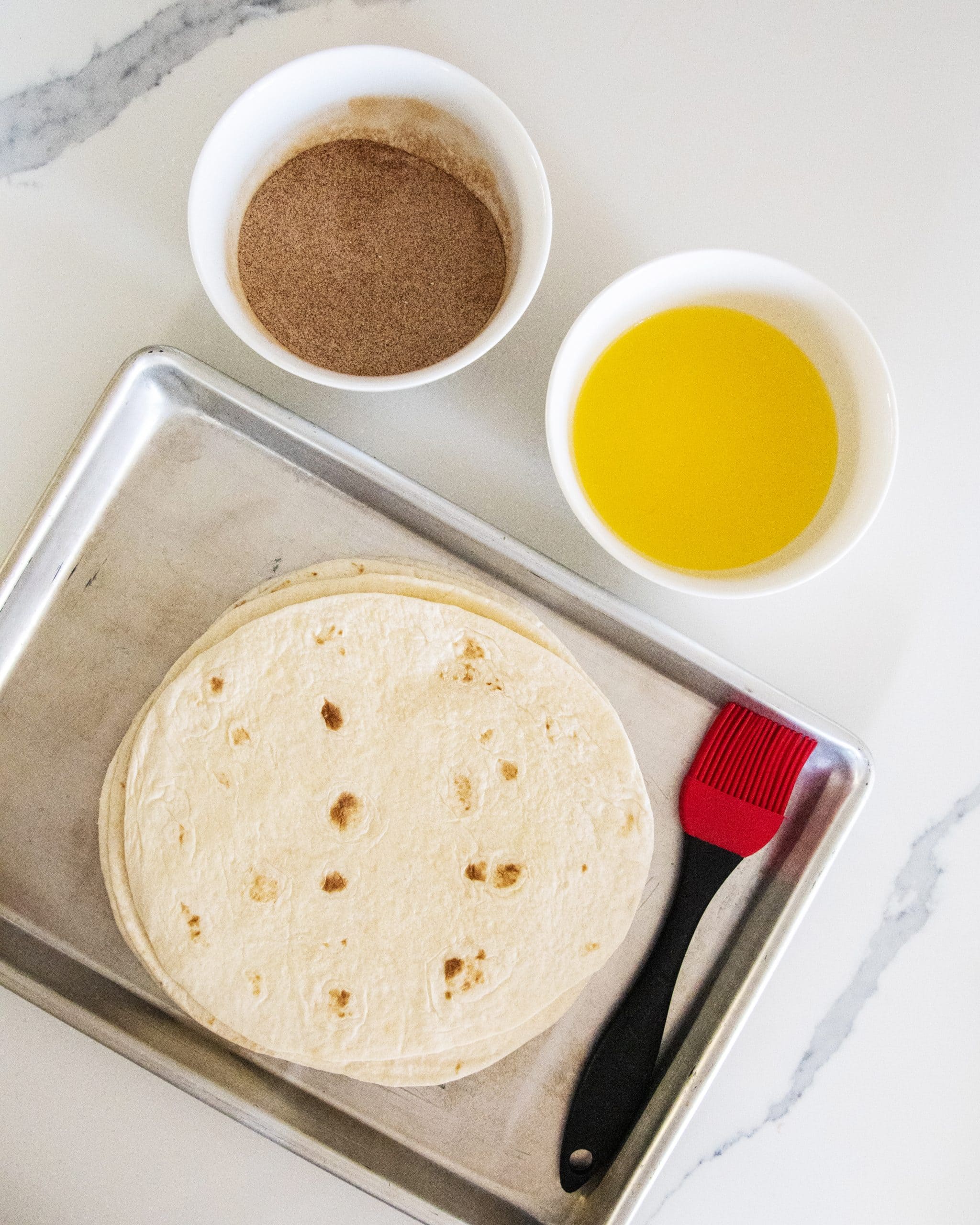 A bowl of cinnamon sugar, a bowl of melted butter, and tortillas on a baking pan.