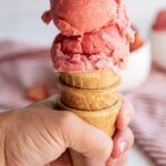 A hand holding an ice cream cone with strawberry sherbet in it.