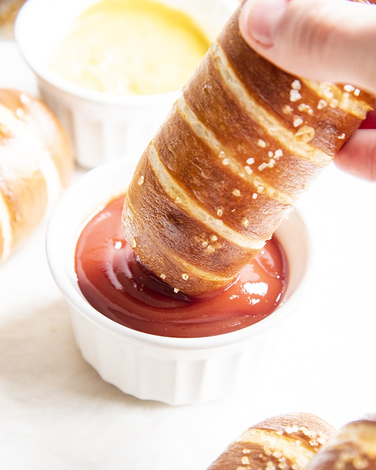 A pretzel wrapped hot dog being dipped in ketchup
