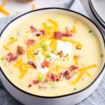 A close up of a bowl of baked potato soup. The soup is yellow colored, and topped with shredded cheese, and crumbled bacon.