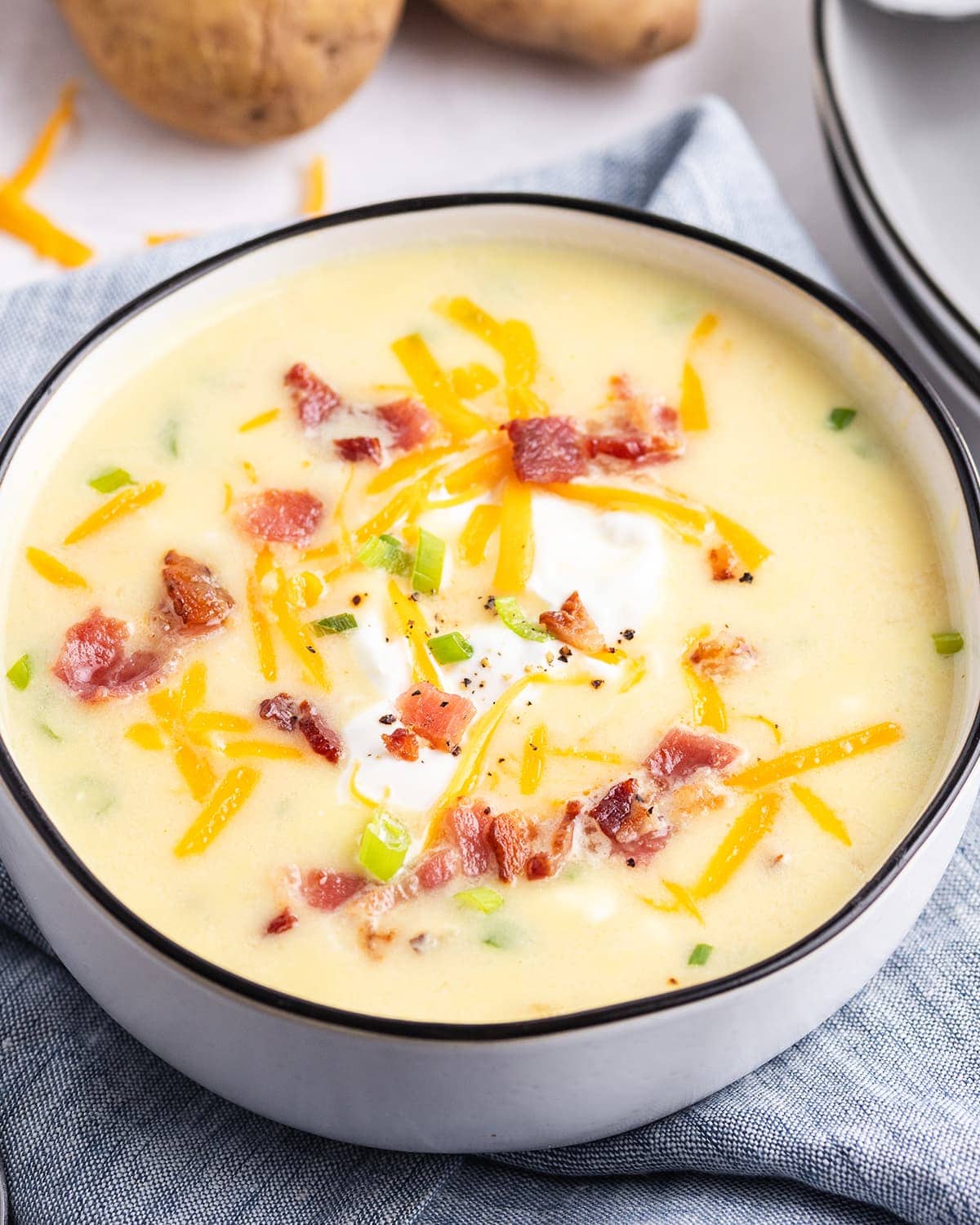 A close up of a bowl of baked potato soup. The soup is yellow colored, and topped with shredded cheese, and crumbled bacon.