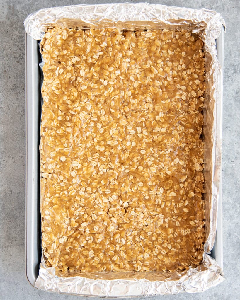 Oatmeal cookie dough pressed into a baking pan that is lined with foil.