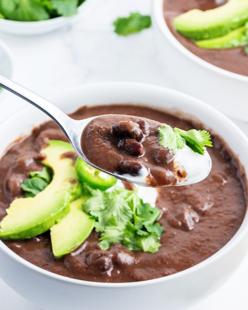 A spoonful of black bean soup above a bowl of the soup topped with avocado slices.