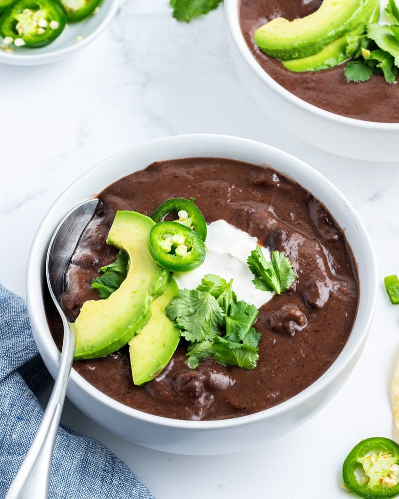 A bowl of creamy black bean soup topped with avocado slices, jalapenos, and sour cream.