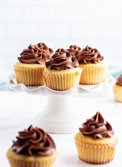 Yellow cupcakes on a white cake stand, each swirled with chocolate buttercream frosting on top.