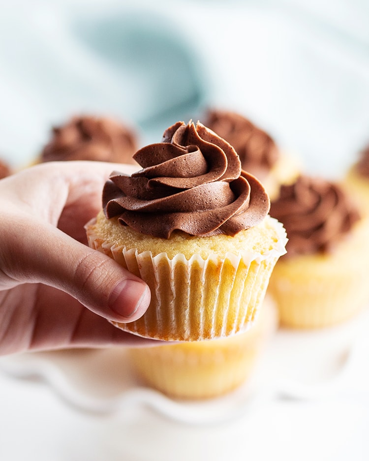 A hand holding a yellow cupcake with chocolate frosting on top.
