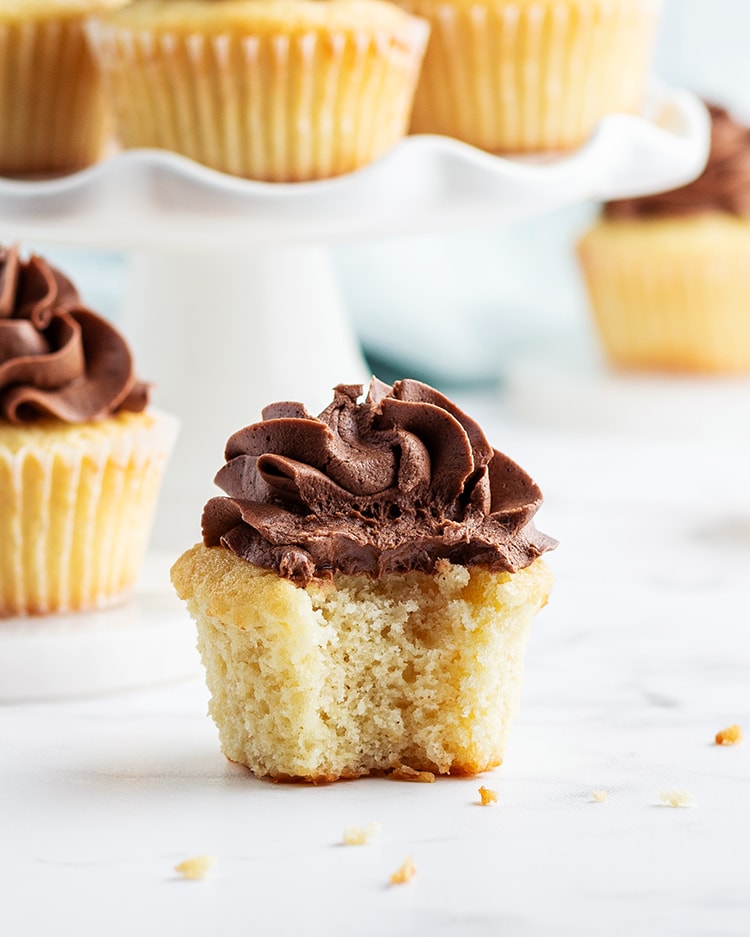 A classic yellow cupcake with chocolate frosting on top, with a bite taken out of it, showing the inside of the cupcake.
