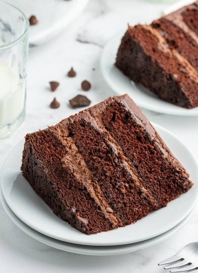 A slice of chocolate cake with three layers and chocolate frosting on a white plate. There is another slice in the back.