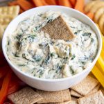 A white bowl full of cheesy spinach dip, with a wheat thin cracker in the middle.