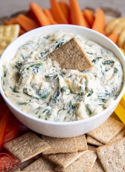 A white bowl full of cheesy spinach dip, with a wheat thin cracker in the middle.