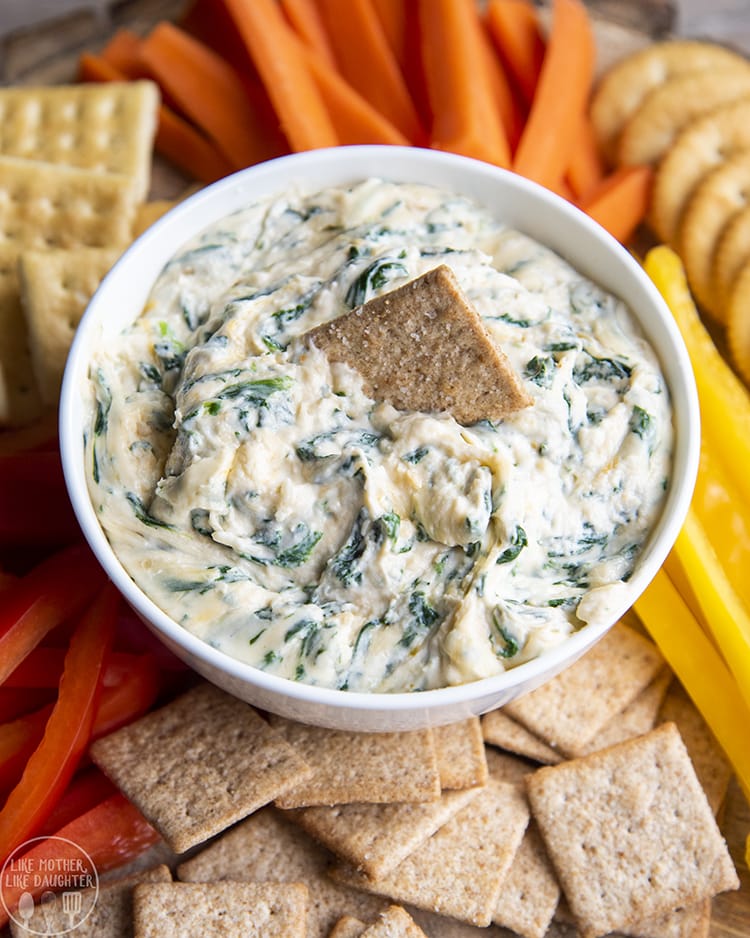 A bowl of a cheesy spinach dip, surrounded by crackers, and carrots, with a wheat thin in the dip.