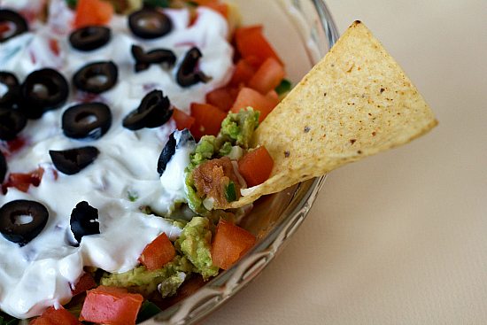 Close up of a chip dipping into a glass bowl full of seven layer dip.