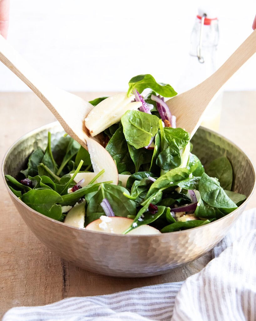 Two wooden spoons lifting and tossing a spinach salad in a bowl.