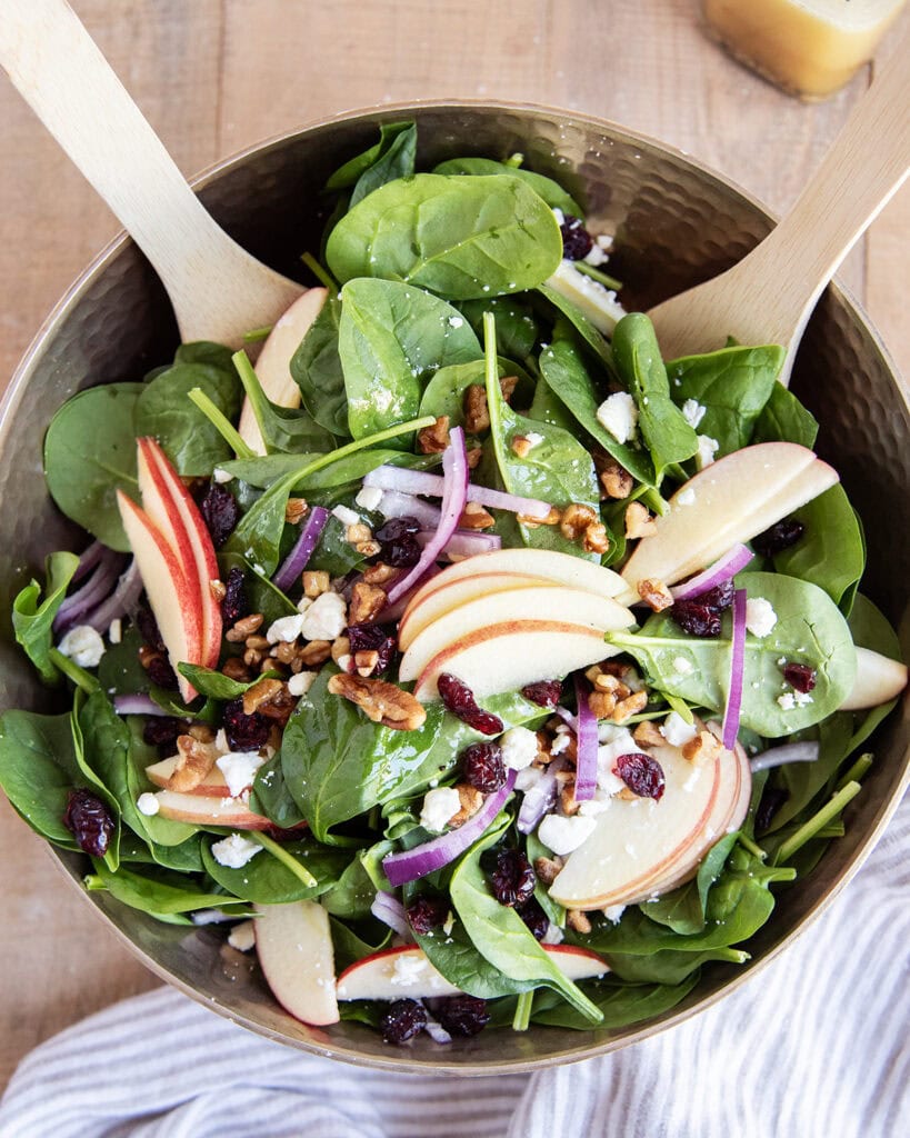 An overhead photo of a golden bowl full of spinach salad with apple slices, and red onion slices.
