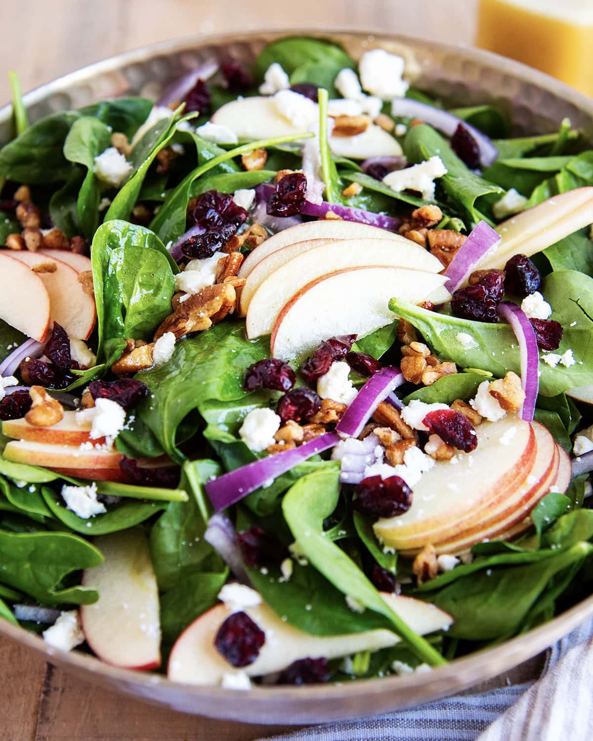 A bowl of spinach salad with apple slices, red onion, cranberries, and feta.