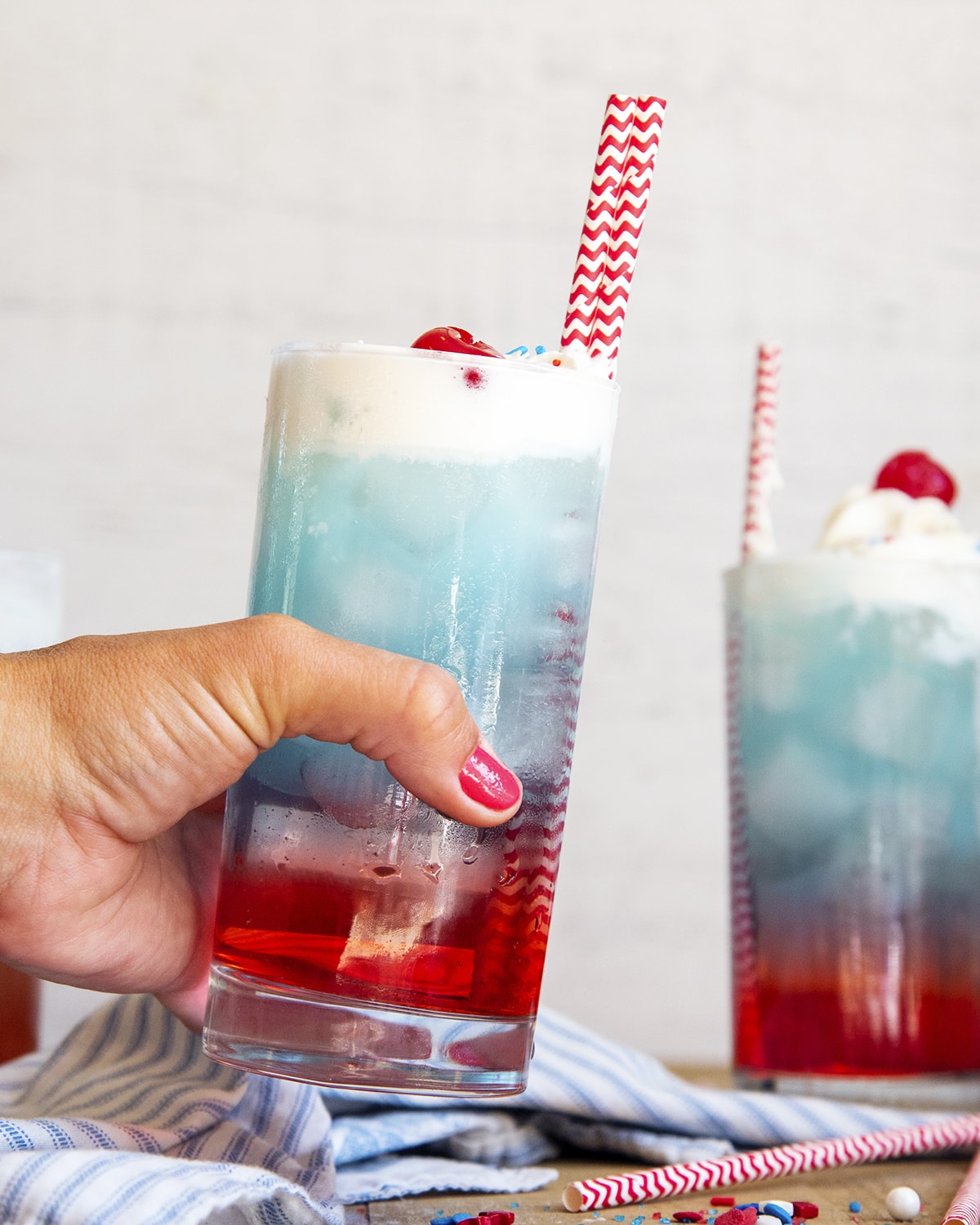 A hand holding a glass of a drink that is red on bottom, blue on top, and topped with whipped cream.