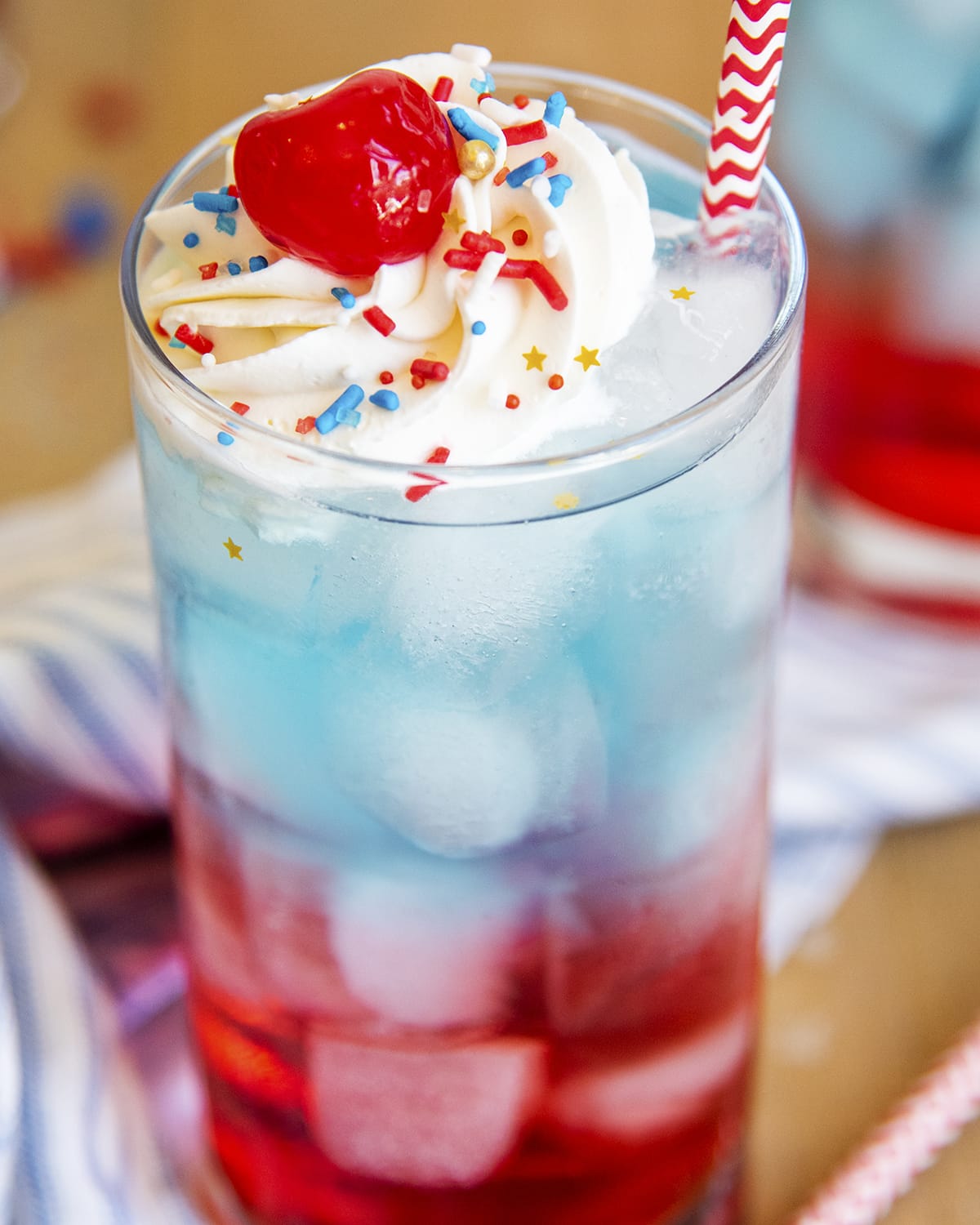 A close up of a glass full of a red and blue drink topped with whipped cream and a cherry.