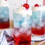 Three glasses of 4th of July Layered Drinks with red bottoms, blue tops, and topped with whipped cream and a cherry.