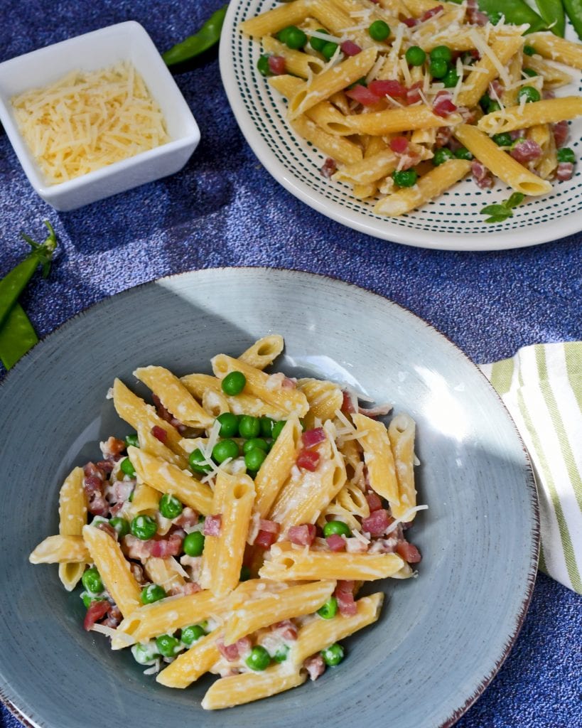 Two plates of penne pasta in a cream sauce with ham and green peas on plates with a small bowl of parmesan cheese on the side