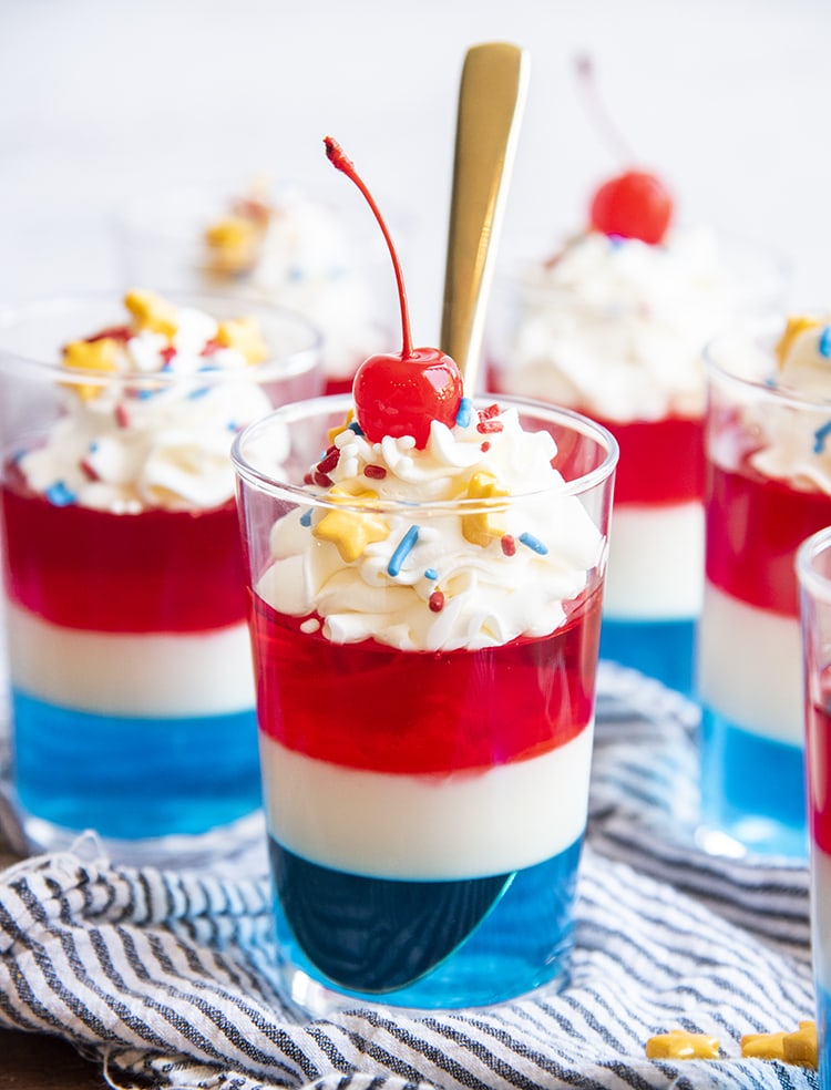 Red White and Blue Jello in a cup topped with whipped cream, and yellow star sprinkles and a maraschino cherry with a spoon in the cup