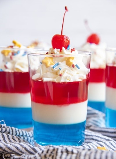 Red White and Blue Jello in a cup topped with whipped cream, and yellow star sprinkles and a maraschino cherry