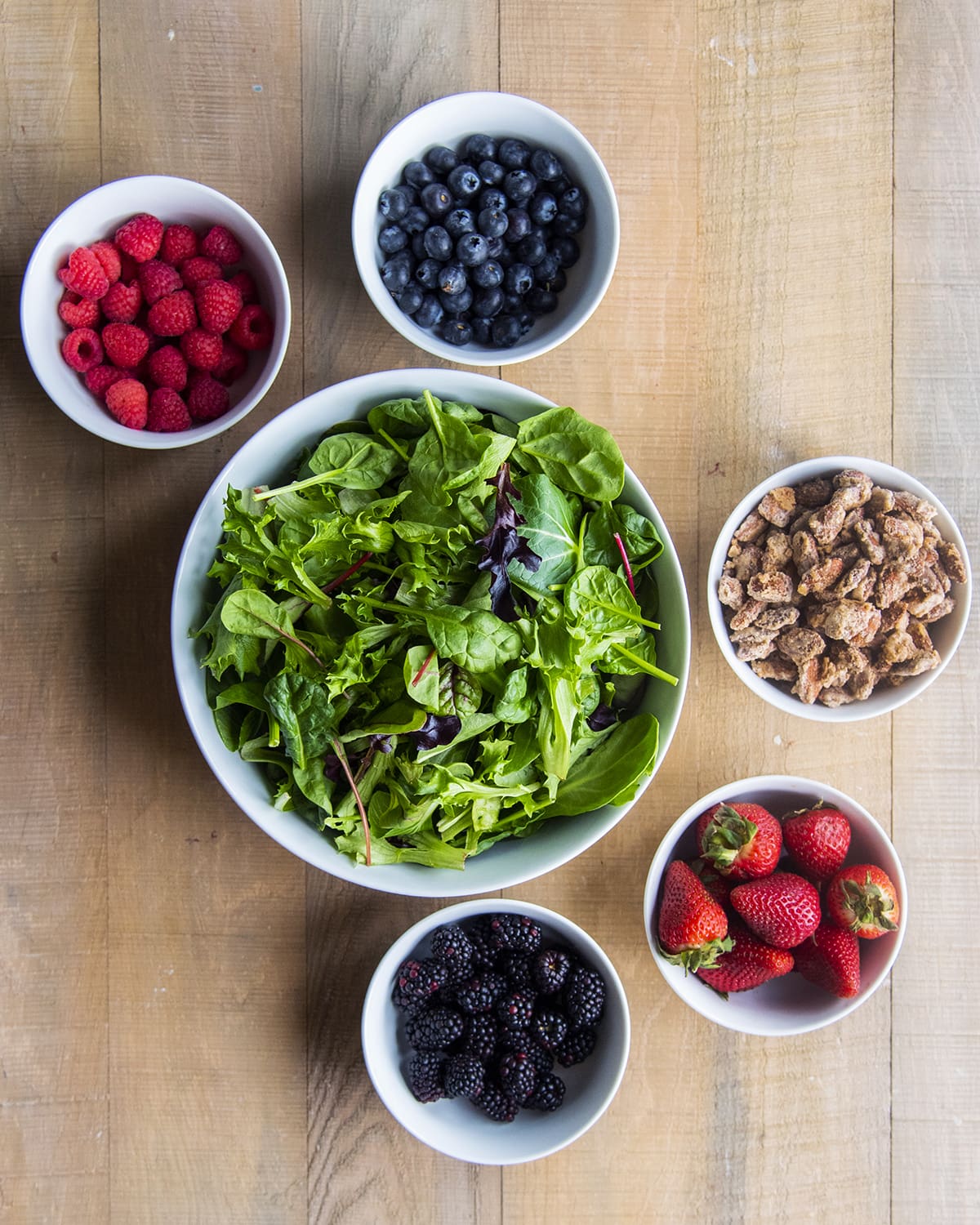 A big bowl of mixed green lettuce with surrounding bowls of strawberries, blueberries, raspberries, blackberries, and candied almonds.