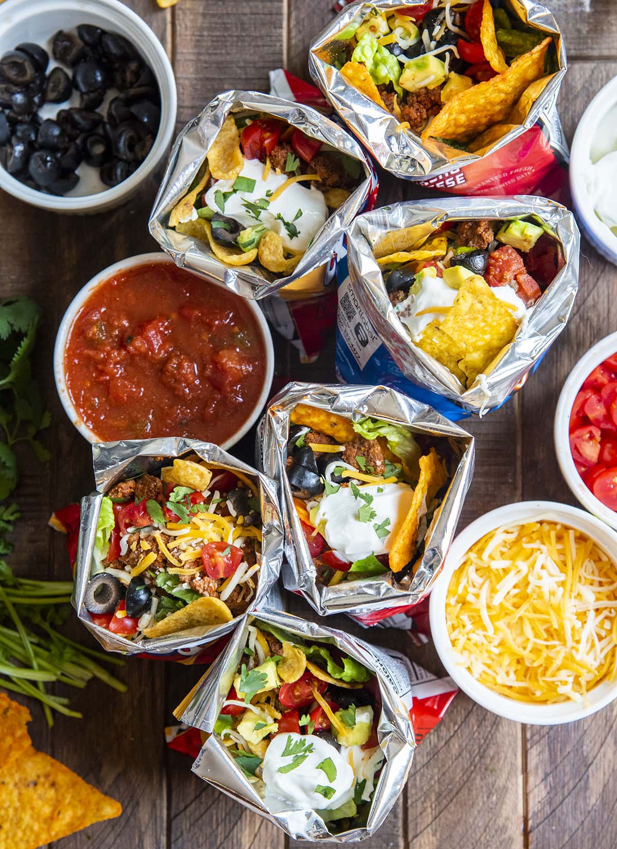 A few walking tacos on a wooden board with toppings in bowls next to them.